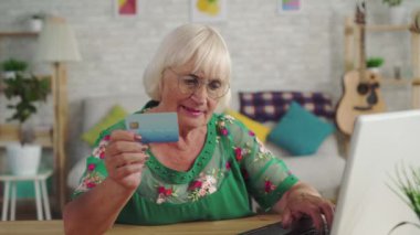 Gözlük gri saçlı yaşlı kadın bir banka kartı tutar ve bir dizüstü bilgisayar kullanır