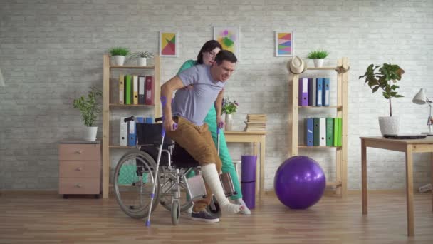 Doctor helps a man get up with a leg injury from a wheelchair after an injury — Stock Video