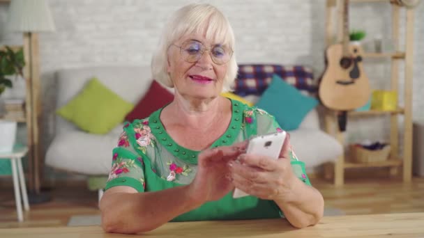 Anciana con pelo gris utiliza un smartphone sentado en una mesa en la sala de estar — Vídeos de Stock