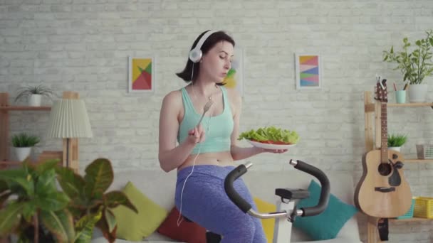 Jovem mulher esportiva em uma bicicleta estacionária ouvindo música e comendo legumes — Vídeo de Stock