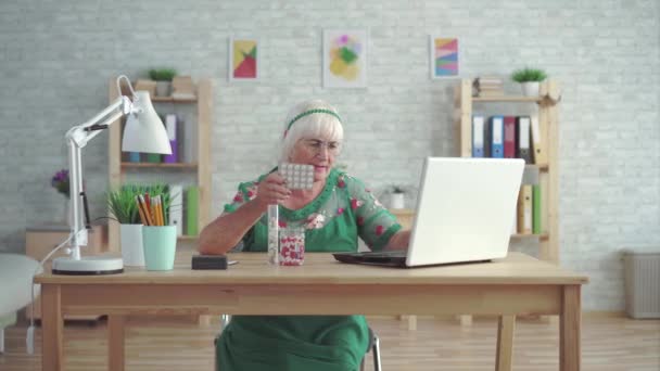 Γριά γυναίκα κοιτάζει σε ένα φορητό υπολογιστή πληροφορίες σχετικά με τα χάπια που κάθονται σε ένα τραπέζι — Αρχείο Βίντεο