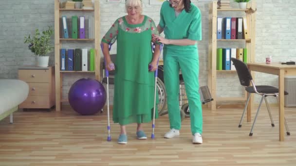 Медсестра в реабилитационной клинике поддерживает пожилую женщину на костылях — стоковое видео
