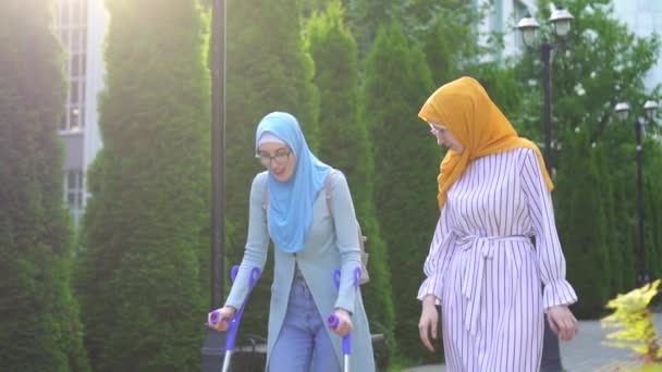 Positivo sonriendo joven mujer en hijab en muletas después de lesión con novia caminando en parque — Vídeos de Stock