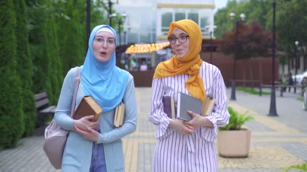 Dos chicas musulmanas sonrientes positivas en hijab y elegantes estudiantes de gafas con libros en sus manos caminando por la calle — Vídeos de Stock