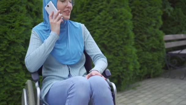 Porträtt ung muslimsk kvinna funktionshindrade i en traditionell halsduk kommunicerar på en smartphone sitter i en rullstol i parken — Stockvideo