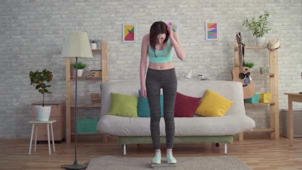 Expressive distressed young woman in sportswear measures her weight on the floor scales — Stock Video