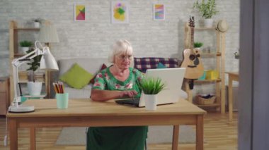 Büyükanne oturma odasında ki masada otururken dizüstü bilgisayarında yazıyor.