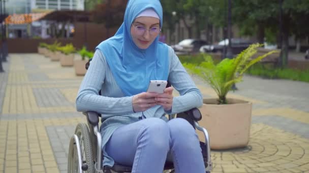 Portret młoda atrakcyjna muzułmańska kobieta niepełnosprawna w tradycyjnym szalik na wózku inwalidzkim używa smartfona siedzącego w parku — Wideo stockowe