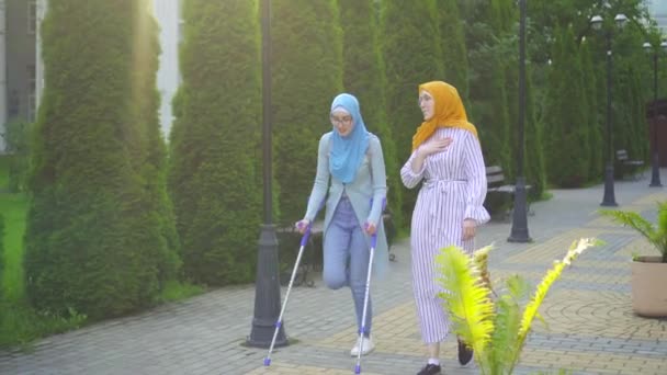 Wanita muslim dengan kaki patah pada kruk berkomunikasi dengan wanita muslim lain dalam syal tradisional di taman — Stok Video