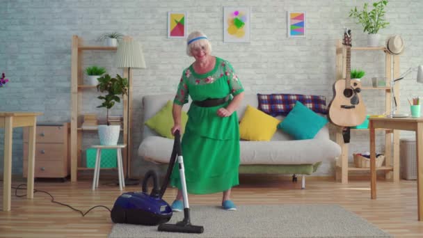 Cheerful happy old woman with gray hair vacuuming the carpet in the living room and dancing — Stock Video