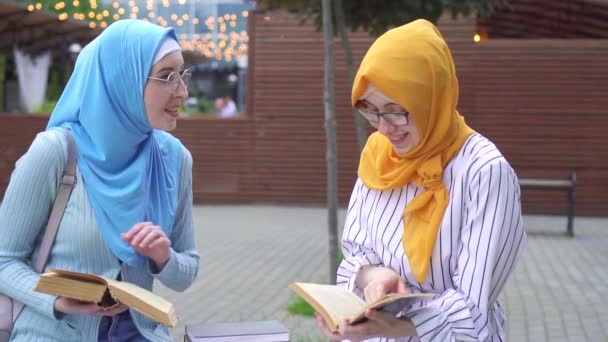 Dos chicas musulmanas sonrientes positivas en hijab y elegantes estudiantes de gafas leyendo libros sentados en un banco — Vídeos de Stock