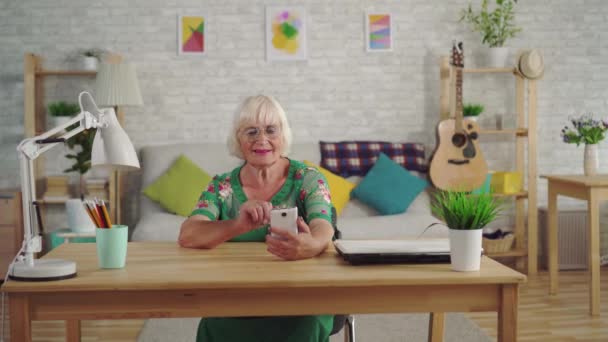 Mujer pensionista con el pelo gris utiliza un teléfono inteligente sentado a la mesa en la sala de estar — Vídeo de stock