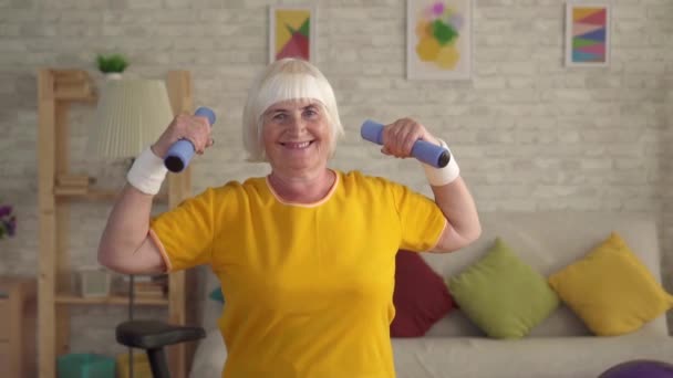 Старшая фитнес-старая женщина позирует с гантелями и смотрит в камеру улыбаясь — стоковое видео