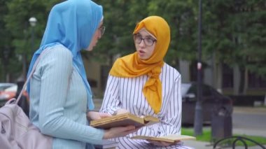Ellerinde kitap olan geleneksel eşarplı iki Müslüman kadın öğrenci parkta iletişim kuruyor