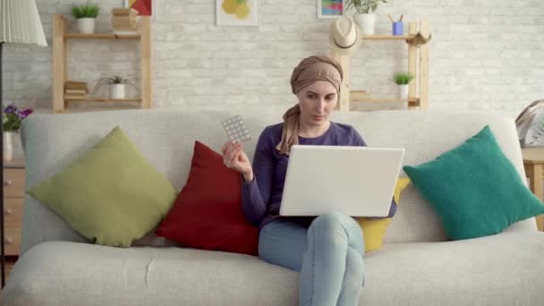 Mujer joven con cáncer en una bufanda después de la quimioterapia con pastillas en la mano utiliza una computadora portátil en busca de información en Internet — Vídeos de Stock