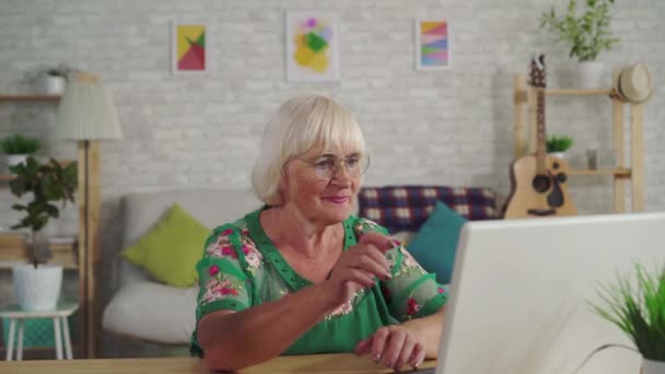 Задумчивая пожилая женщина находит решение и использует ноутбук, сидя за столом в гостиной — стоковое видео