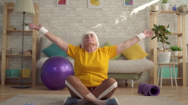 Mujer mayor activa positiva haciendo yoga en la alfombra en la sala de estar — Vídeos de Stock