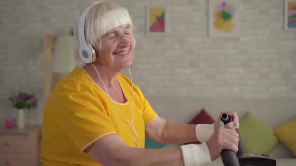 Активная старуха в наушниках занимается кардиотренировкой на велотренажере вблизи — стоковое видео
