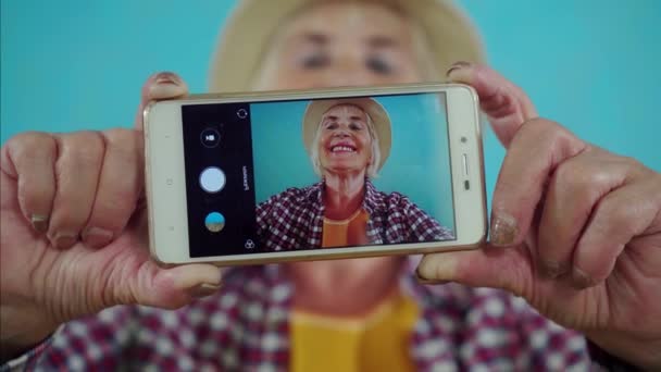 Positivo anziana donna sorridente si spara con un telefono fotocamera in piedi su uno sfondo blu, la vista attraverso la fotocamera — Video Stock