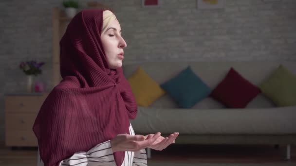 Μουσουλμανική γυναίκα σε ένα παραδοσιακό κασκόλ προσεύχεται στο χαλί στο σαλόνι της κοντά — Αρχείο Βίντεο