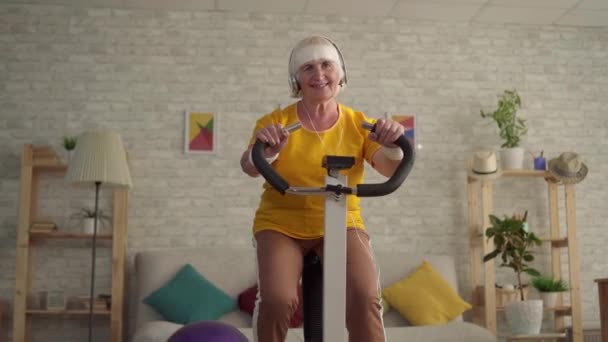 Actieve oude vrouw in hoofdtelefoon bezig met cardiotraining op een hometrainer — Stockvideo