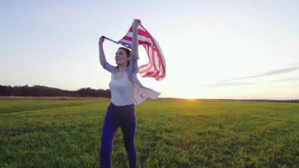 Giovane donna positiva corre con una bandiera USA su un campo al tramonto — Video Stock