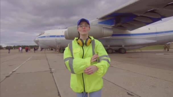 Работница аэропорта на фоне самолета позирует скрещивая руки улыбаясь и глядя в камеру — стоковое видео