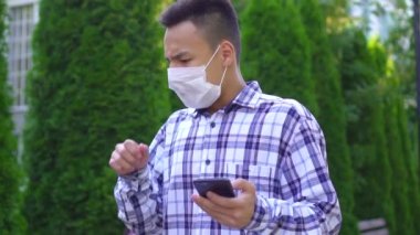 yüzünde tıbbi maske genç asyalı adam parkta öksürük geçer ve bir akıllı telefon yavaş mo kullanır