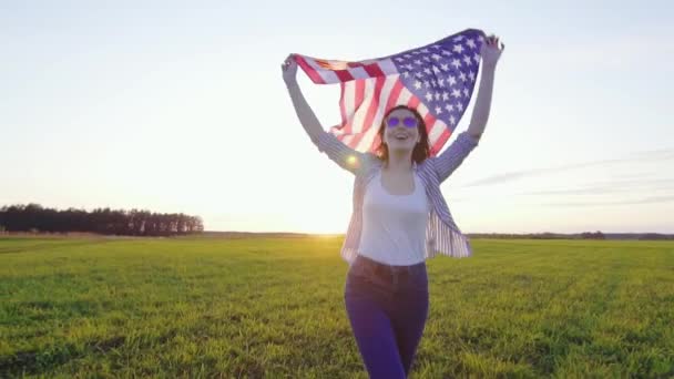 Jovem mulher positiva com uma bandeira dos EUA corre através de um campo ao pôr do sol slow mo — Vídeo de Stock