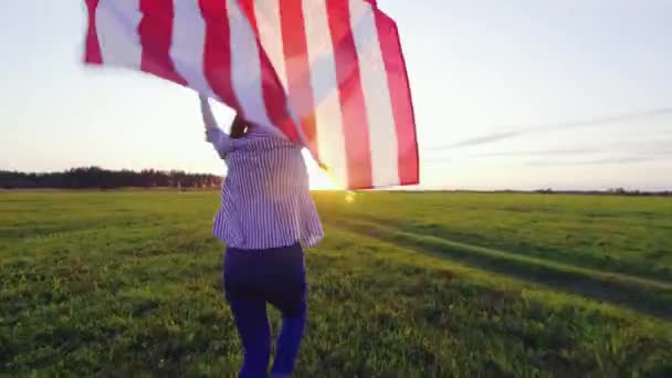 Giovane donna corre con una bandiera USA su un campo al tramonto vista posteriore — Video Stock