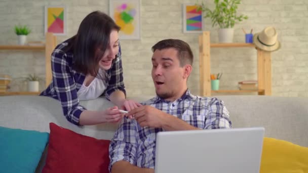 Jonge positieve vrouw dansen met vreugde dat ze zwanger is, verdrietig man in shock zittend op de laptop op de Bank en omhoog — Stockvideo