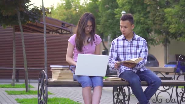 Mladí asijští studenti s knihou a ženou s notebookem, který sedí na lavičce. — Stock video