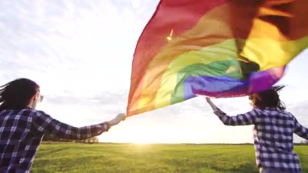 Δύο νεαρά κορίτσια λεσβίες με πουκάμισα και γυαλιά τρέχουν κατά μήκος του γηπέδου στο ηλιοβασίλεμα με σημαία ΛΟΑΤ — Αρχείο Βίντεο