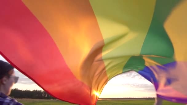 Duas jovens mulheres feministas jovens e alegres em camisas correm pelo campo com uma bandeira LGBT slow mo — Vídeo de Stock