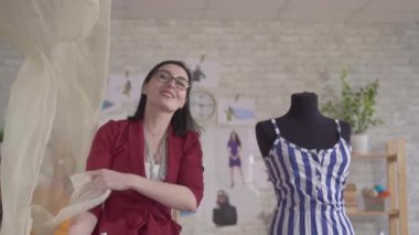 Genç, tatlı kadın moda tasarımcısı, gözlüklü, kumaş yavaş çekimde çalışıyor.