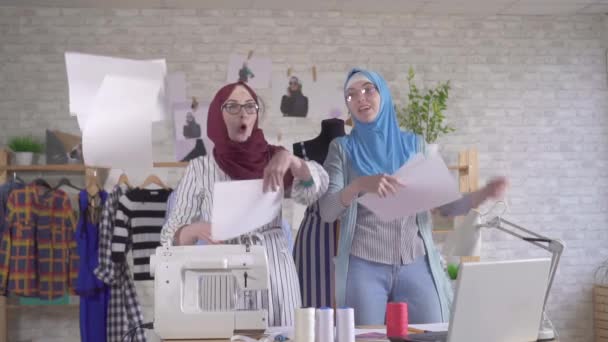 Две позитивные молодые мусульманки-модельеры в национальных платках швыряют бумажки для удовольствия медленно — стоковое видео
