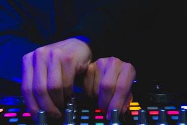DJ müzik çalıyor. Ses karıştırıcı kontrolörü, düğmeleri ve sürgüleri var. Karanlığın içinde parlayan kontrollü pikaplarla karıştırma güvertesinde.