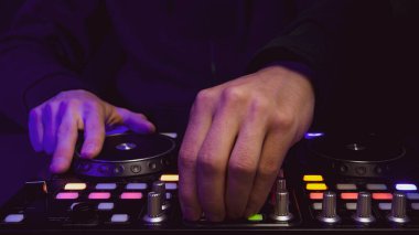 DJ müzik çalıyor. Ses karıştırıcı kontrolörü, düğmeleri ve sürgüleri var. Karanlığın içinde parlayan kontrollü pikaplarla karıştırma güvertesinde.