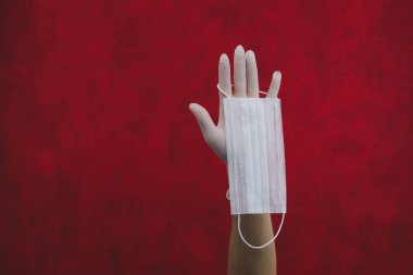 Beyaz tıbbi maske beyaz bir ameliyat eldiveniyle elinde asılı duruyor. Solunum hastalıklarına ve virüse karşı önleyici önlemler. Kopyalama alanı olan kırmızı arkaplan