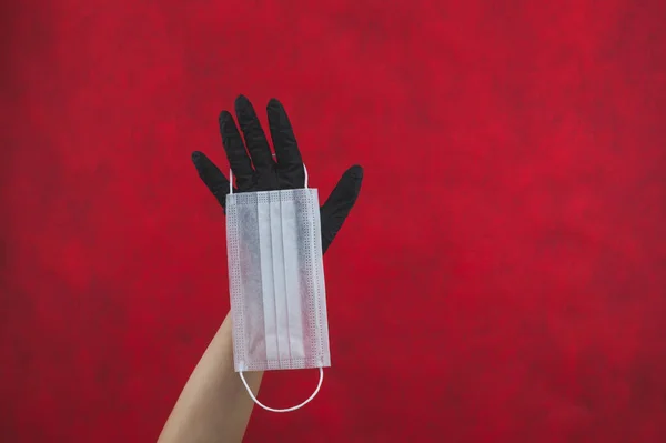 白色的外科口罩挂在手上 戴着一只黑色的医疗手套 预防呼吸道疾病和病毒的预防措施 一次性保护面具 带有复制空间的红色背景 — 图库照片