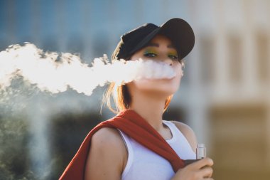 Genç ve güzel hipster Asyalı siyah şapka vape ecig, gün batımında vaping aygıt. Tonlu görüntü. Portre.