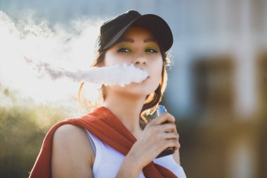 Genç ve güzel hipster Asyalı siyah şapka vape ecig, gün batımında vaping aygıt. Tonlu görüntü. Portre.