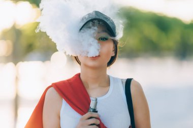 Genç ve güzel hipster Asyalı siyah şapka vape ecig, gün batımında vaping aygıt. Tonlu görüntü. Portre.
