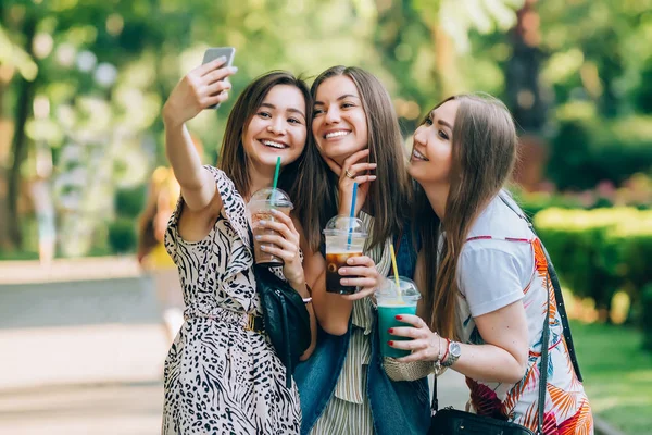 Happy friends in the park on a sunny day . Summer lifestyle portrait of three multiracial women enjoy nice day, holding glasses of milkshakes. Taking picture on mobile phone. Best friends girls having