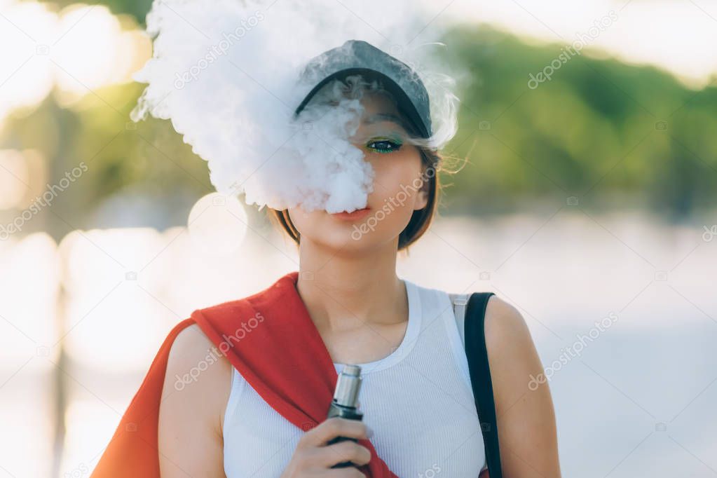 Pretty young hipster asian woman in black hat vape ecig, vaping device at the sunset. Toned image. Closeup.