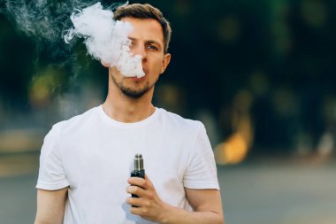 Bir sakal eğlenceli Vaporizers açık olan modern genç adam. Erkek, beyaz t-shirt vape ecig. koyu arka plan, akşam gün batımını şehri. tonlu görüntü.