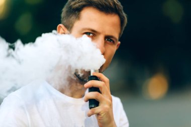 Bir sakal eğlenceli Vaporizers açık olan modern genç adam. Erkek, beyaz t-shirt vape ecig. koyu arka plan, akşam gün batımını şehri. tonlu görüntü.