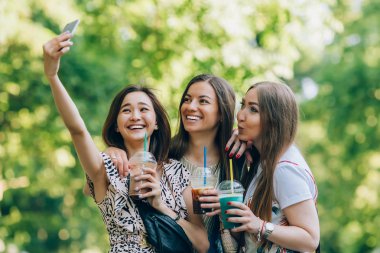 Mutlu arkadaş güneşli bir parkta. Yaz yaşam tarzı üç çok ırklı kadın portresi iyi günler, milkshake bardak tutan tadını çıkarın. Çekici resim üstünde hareket eden telefon. En iyi arkadaşlar kız orgazm