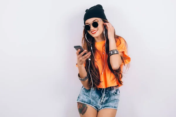 Junge Hipster schöne Frau mit Sonnenbrille, schwarzem Hut und orangefarbenem T-Shirt, die Musik über Kopfhörer in der Nähe der weißen Wand hört, mit einem Handy in der Hand — Stockfoto