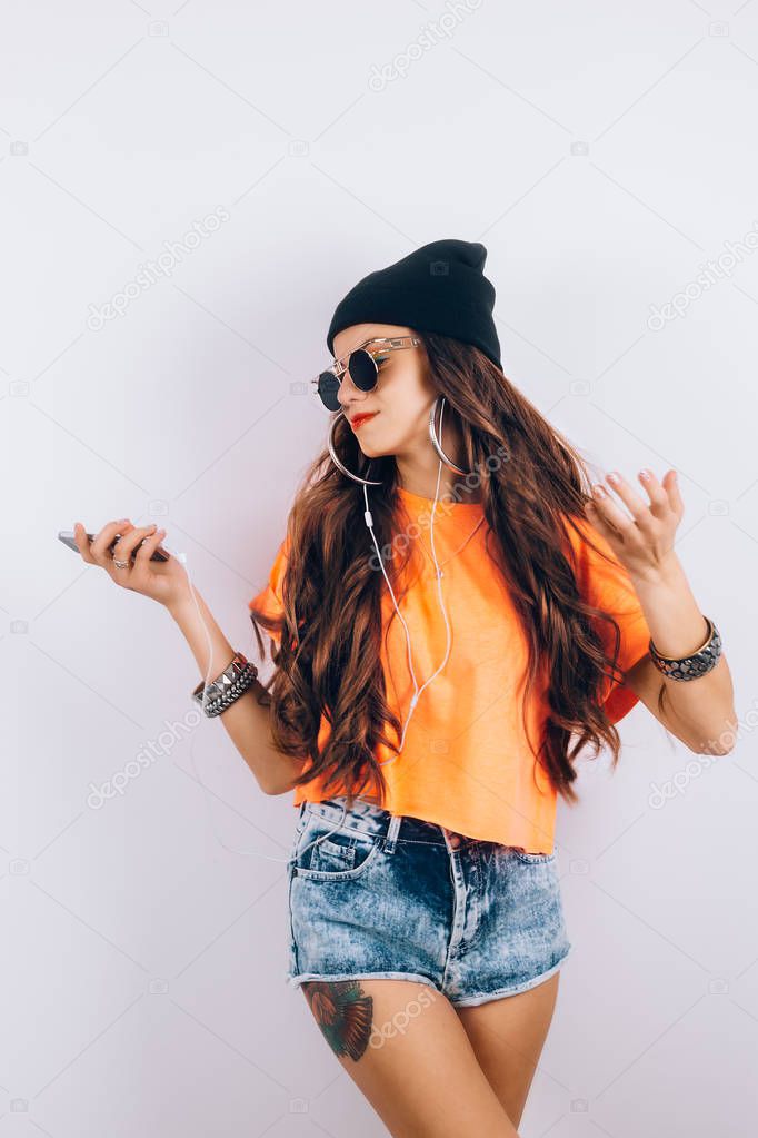 Young hipster beautiful woman in sunglasses wearing in black hat and orange T-shirt listening music in headphones near white wall, holding a cell phone in hand